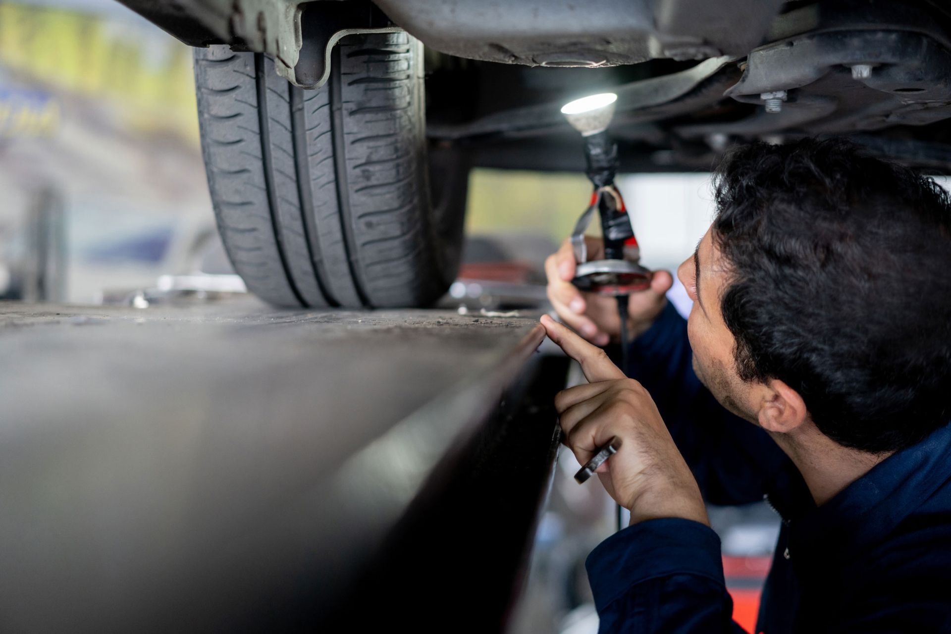 Mechanic checking the alignment of a car — Mildura, VIC — United Panel Works Mechanic inspecting a car's underside with a flashlight, focusing on components near the tire, in a repair shop.
