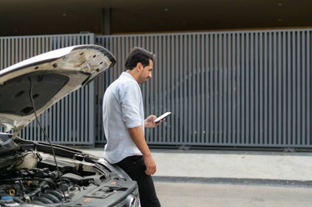Man with car trouble looks at phone beside his open car hood, near a metal gate.