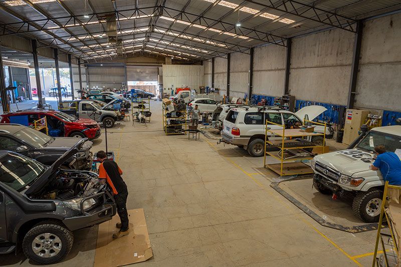 Mechanic shop interior with several vehicles being worked on. Workers in orange and blue.