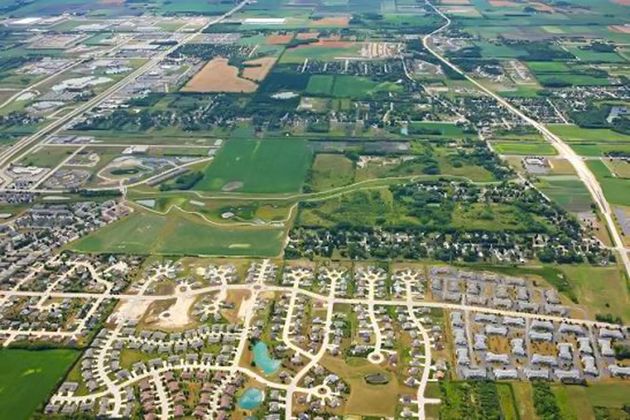 Aerial view of a suburban neighborhood beside farmland, with roads, houses, and green fields