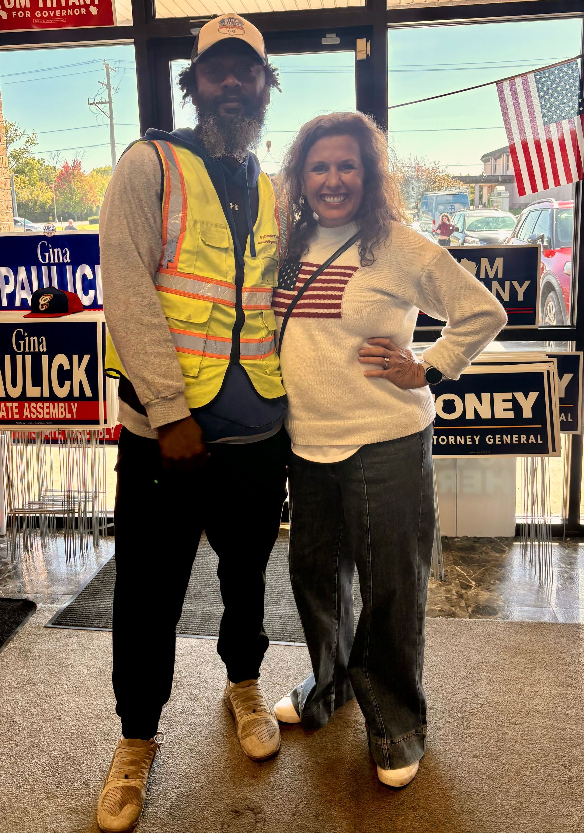 Two people posing together indoors in front of campaign signs, wearing casual clothes and smiling.