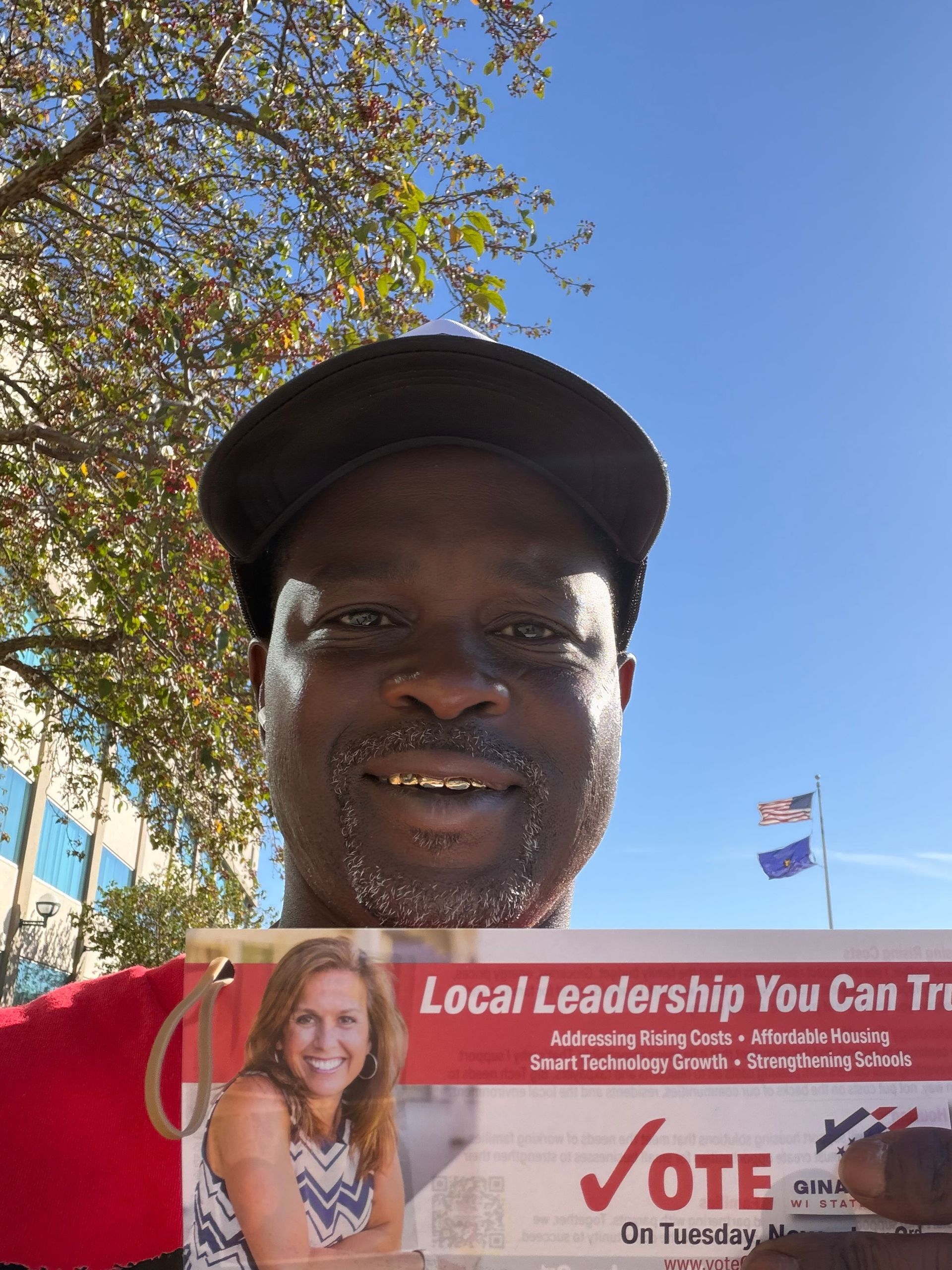 Person holding a red election sign outdoors under a tree and blue sky.