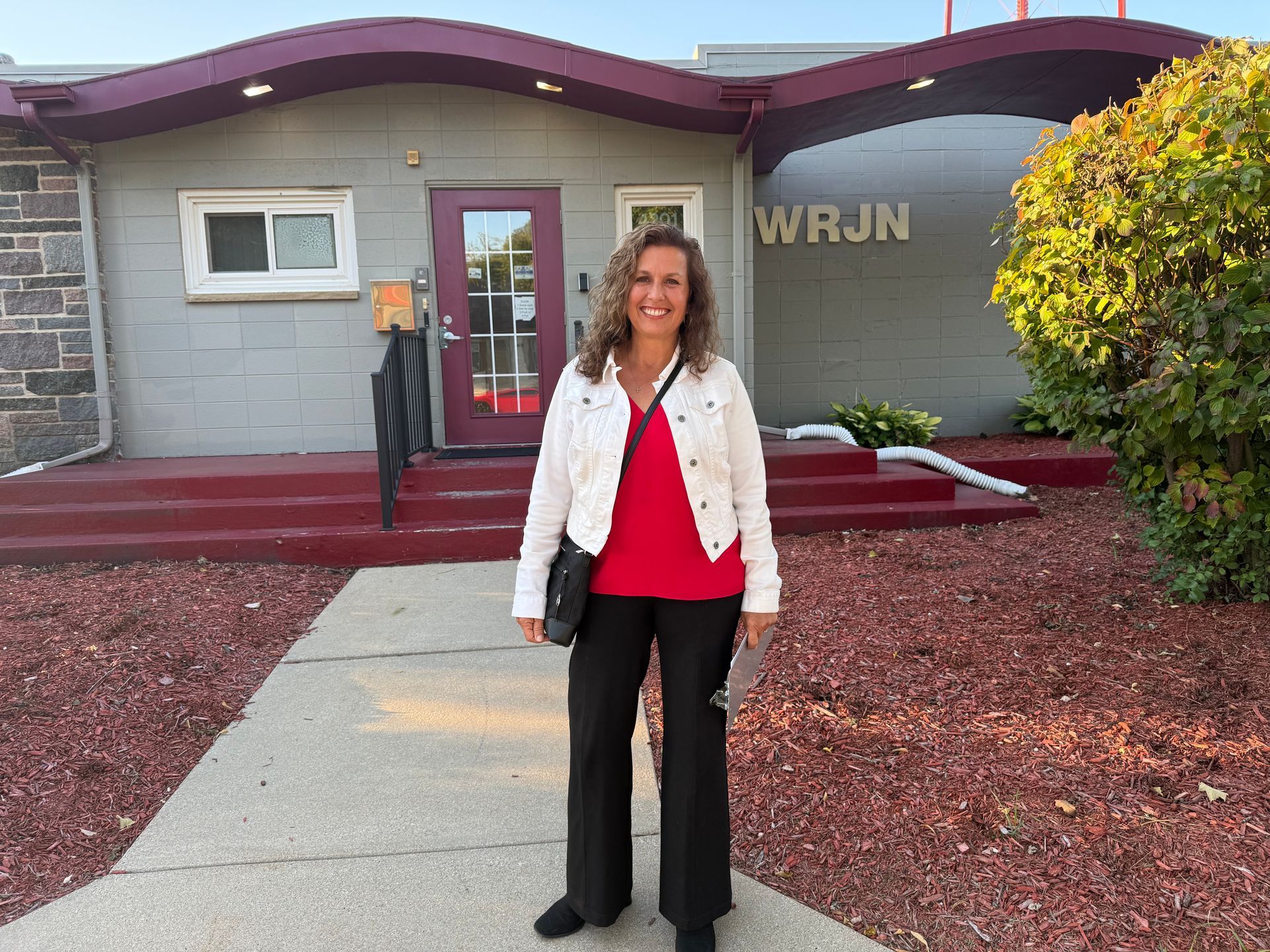Woman standing in front of a building with a WRJN sign, wearing a white jacket and red top