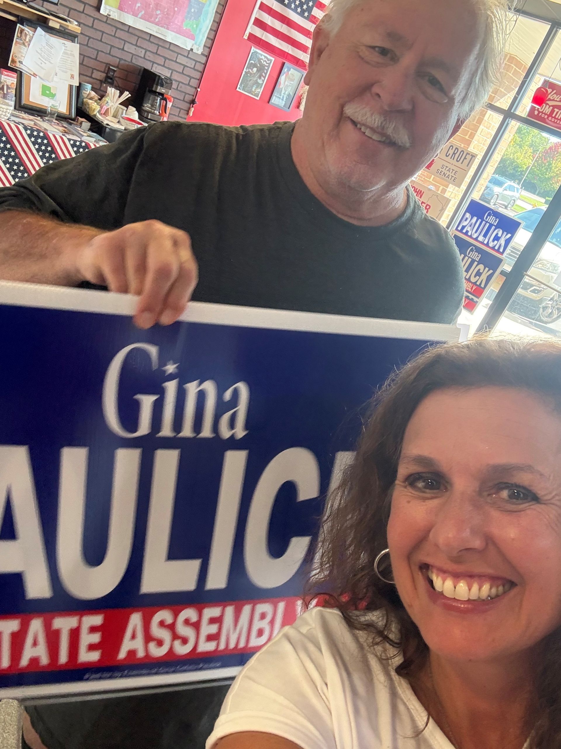 Two smiling people holding a blue political campaign sign indoors