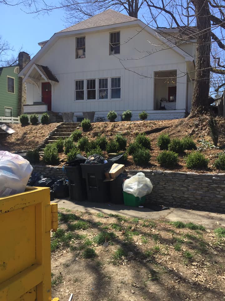 A white house with a red door and a stone retaining wall in front, with a yellow dumpster in the foreground.