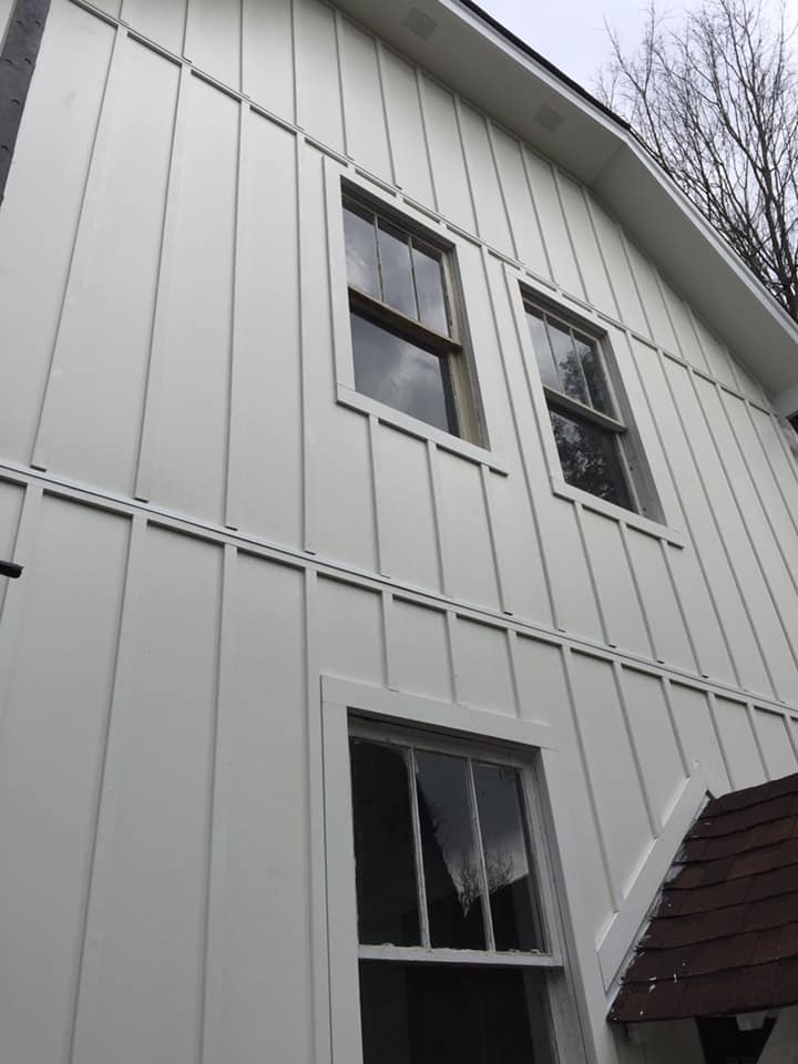 Exterior view of a white, vertical board-and-batten siding wall with three windows and a portion of a brown roof.