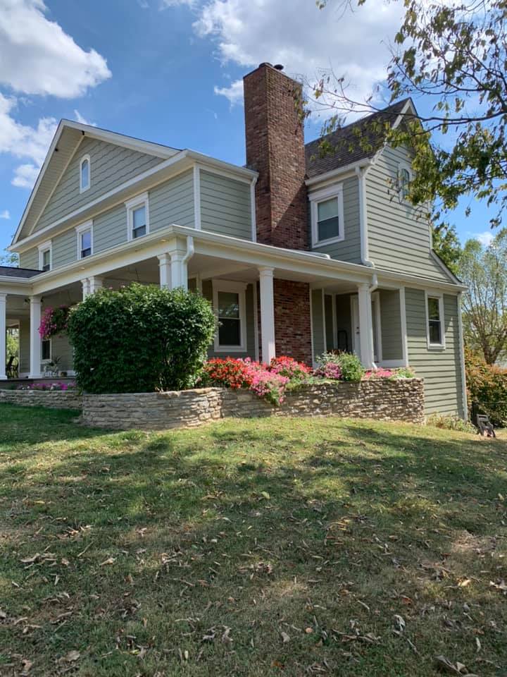 Two-story light green farmhouse with a brick chimney, wrap-around porch, and a tiered stone garden bed with flowers.
