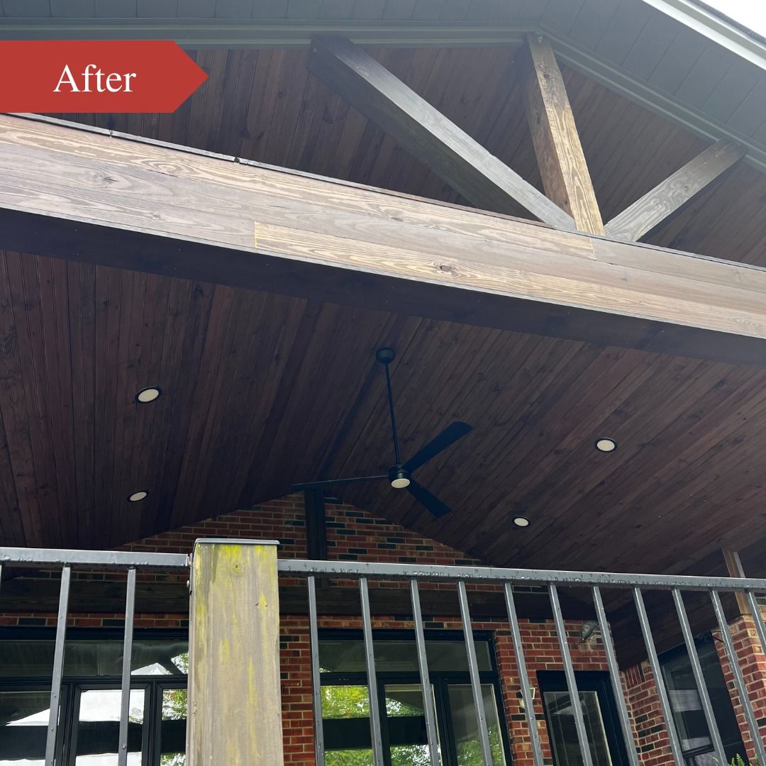 Wooden porch ceiling with exposed beams, railing, and a ceiling fan under a dark roof.
