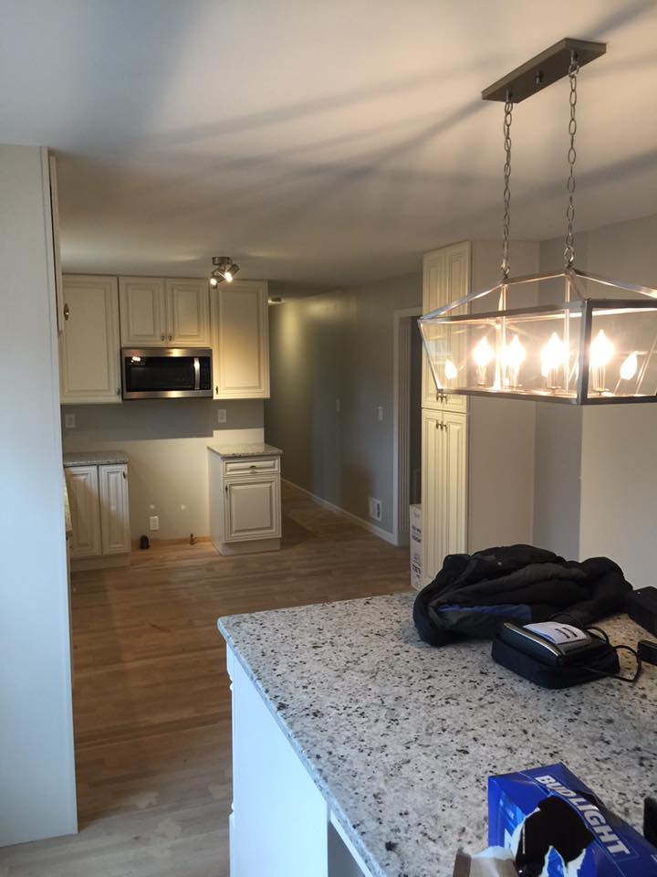 A kitchen interior with white cabinets, a microwave, wood floors, a granite island countertop, and a modern light fixture.