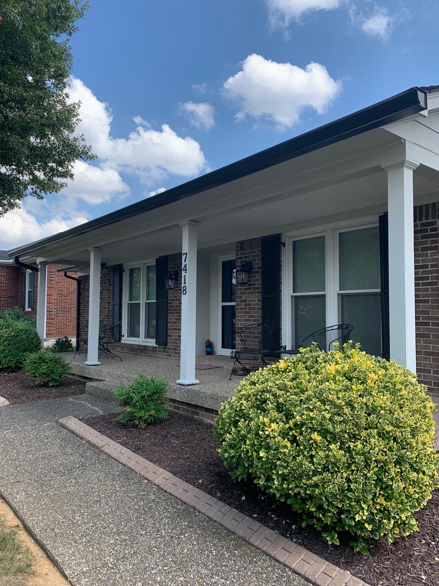 A brick house with a covered front porch, white columns, dark shutters, and a gravel walkway with landscaped bushes.