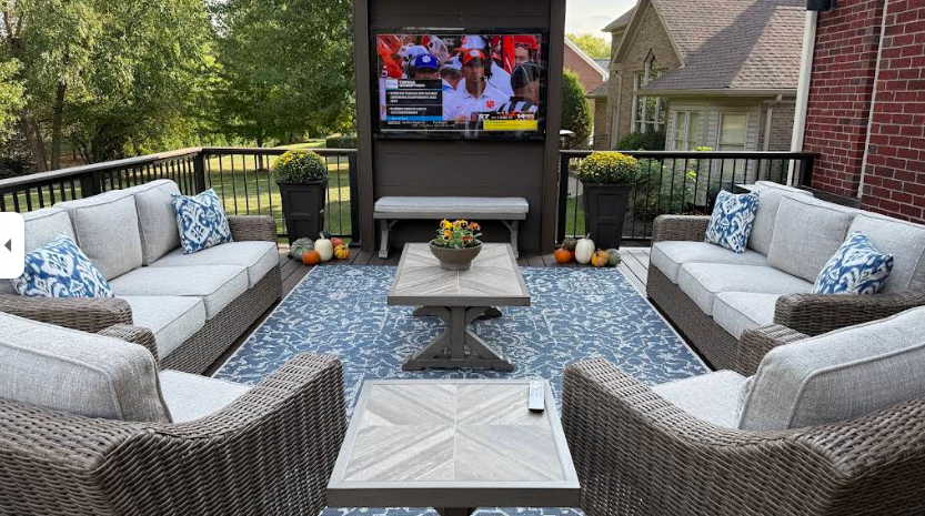 Cozy outdoor patio with wicker seating around a TV and fire table, overlooking a backyard and houses.