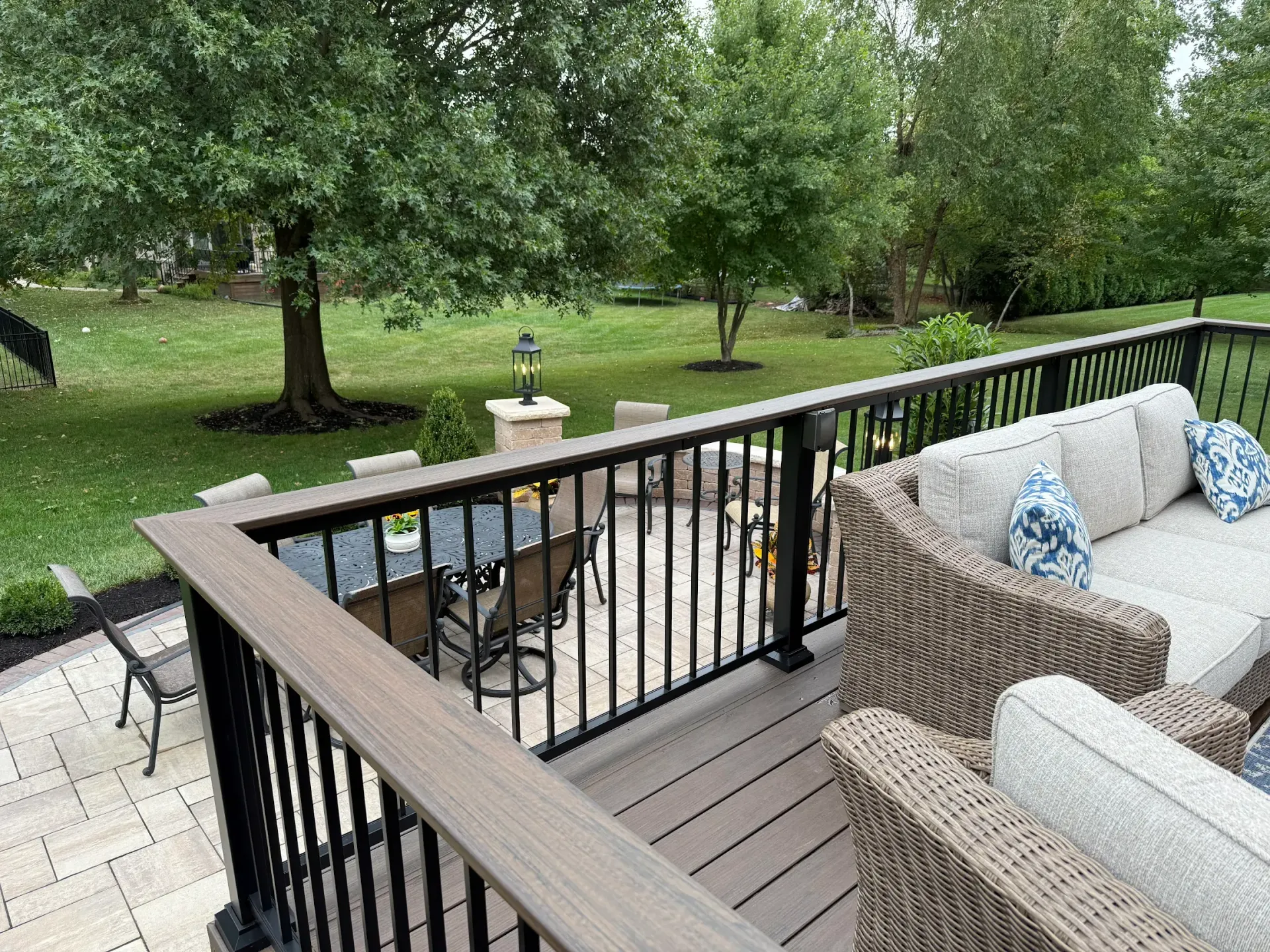 A deck with gray wicker furniture and a black metal railing overlooking a patio, grassy lawn, and mature trees.