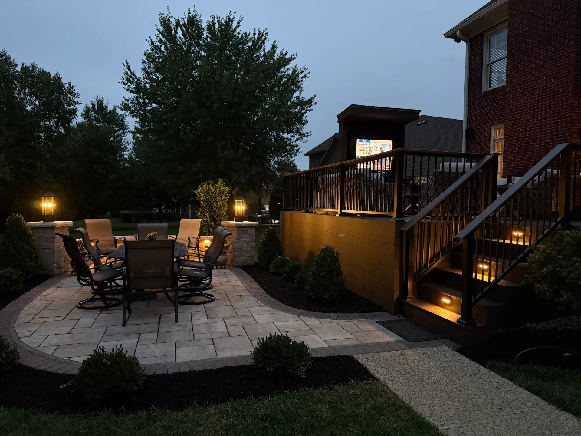 A patio at dusk featuring a dining set, stone flooring, and warm landscape lighting leading to a wooden deck and brick house.