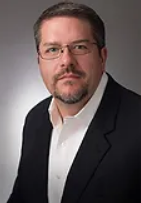 A man with a beard and glasses is wearing a suit and white shirt.