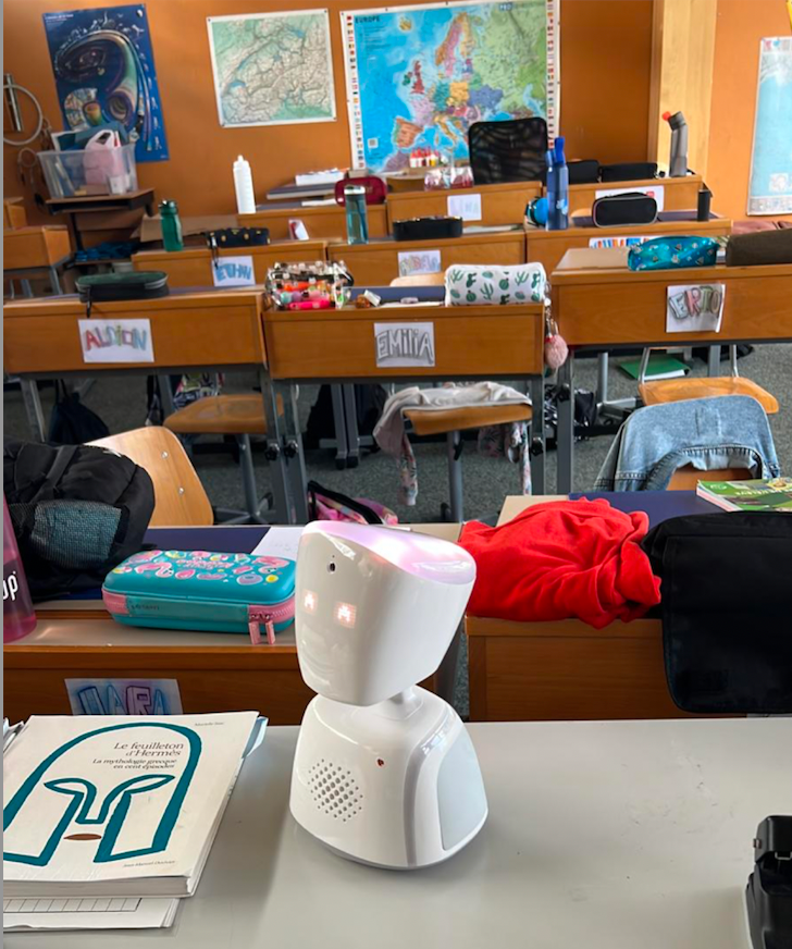 Un robot est assis sur un bureau dans une salle de classe avec un panneau sur le mur qui dit selma