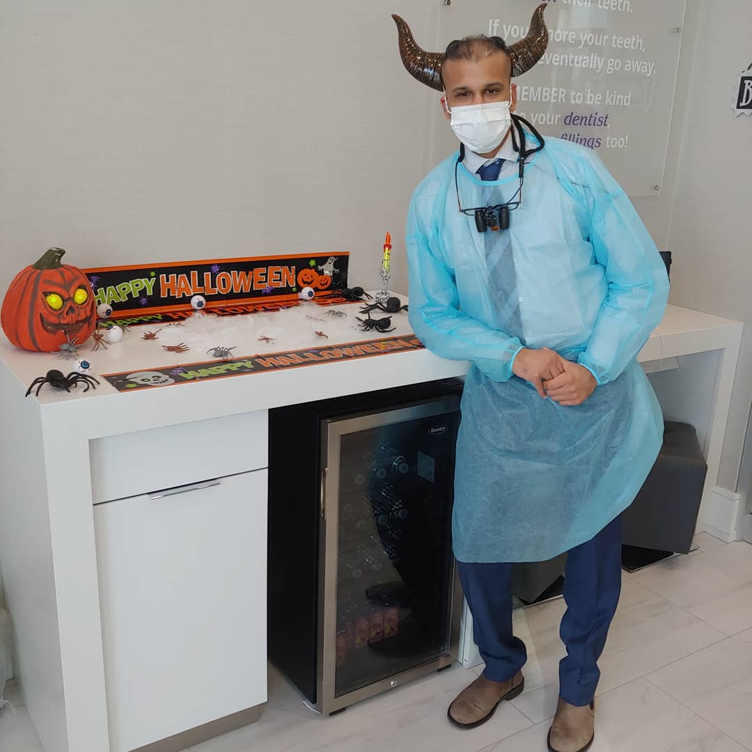 Person in blue protective gown and mask standing beside a Halloween-decorated counter with a pumpkin display.