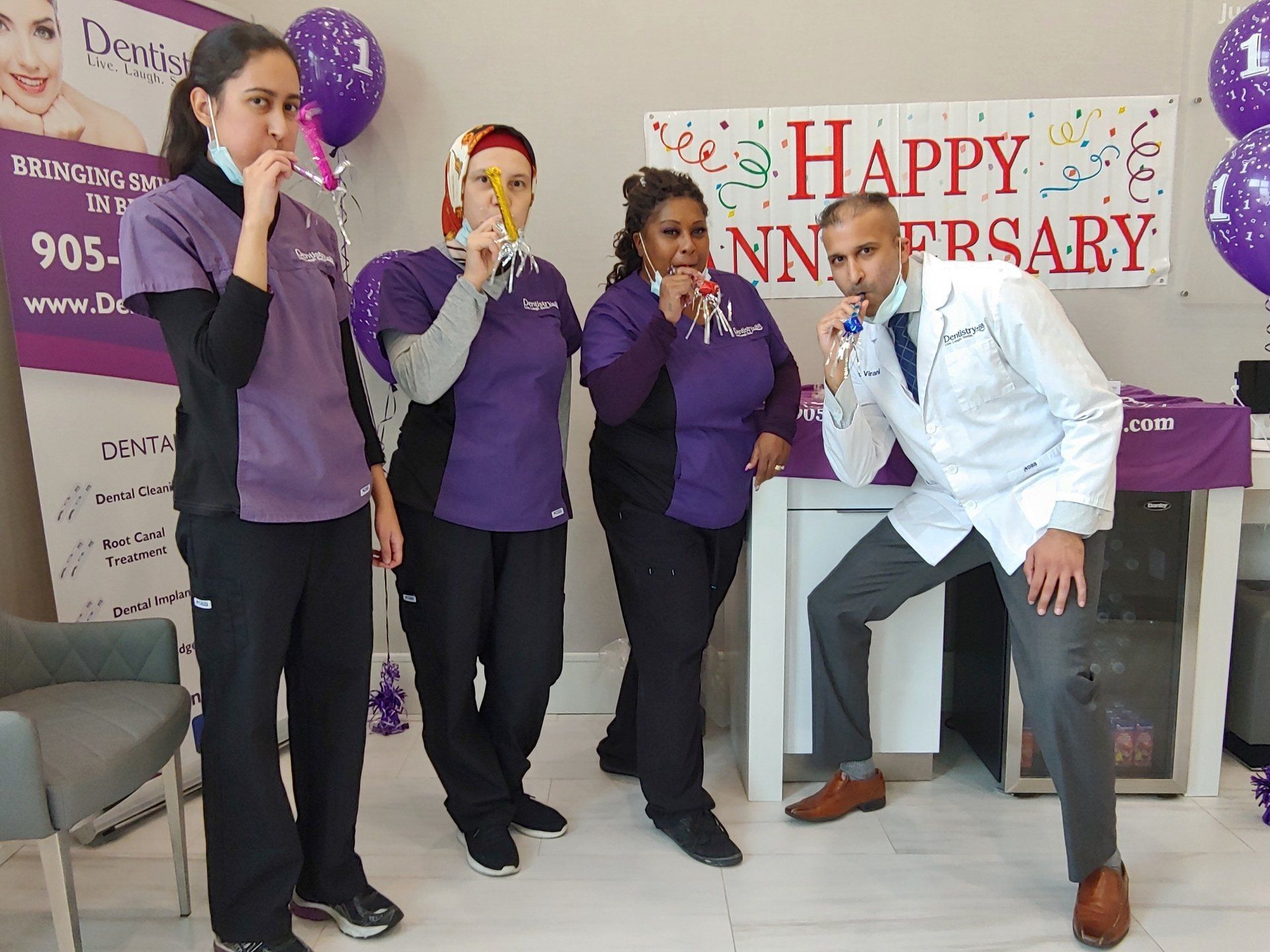 Four people pose at a purple-themed anniversary event with a balloon and “Happy Anniversary” banner.