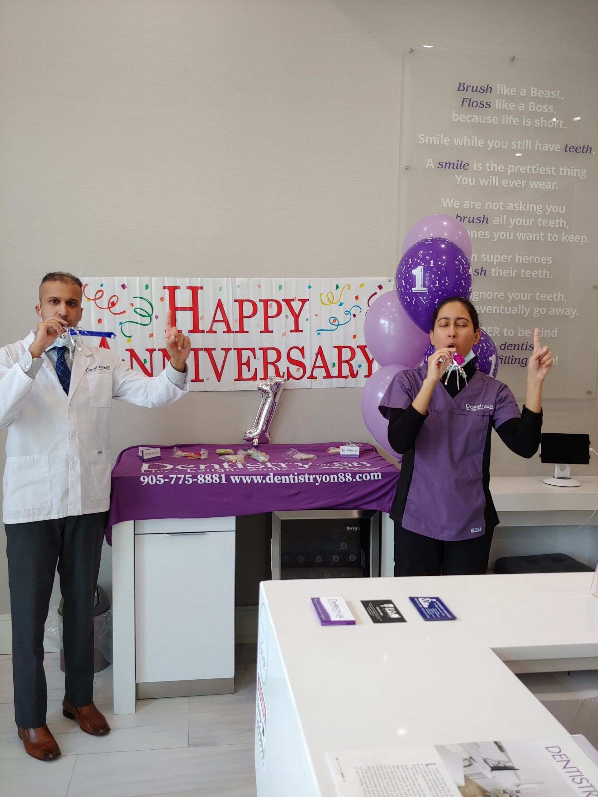 Two people holding microphones at a happy anniversary event with a purple banner and balloon backdrop