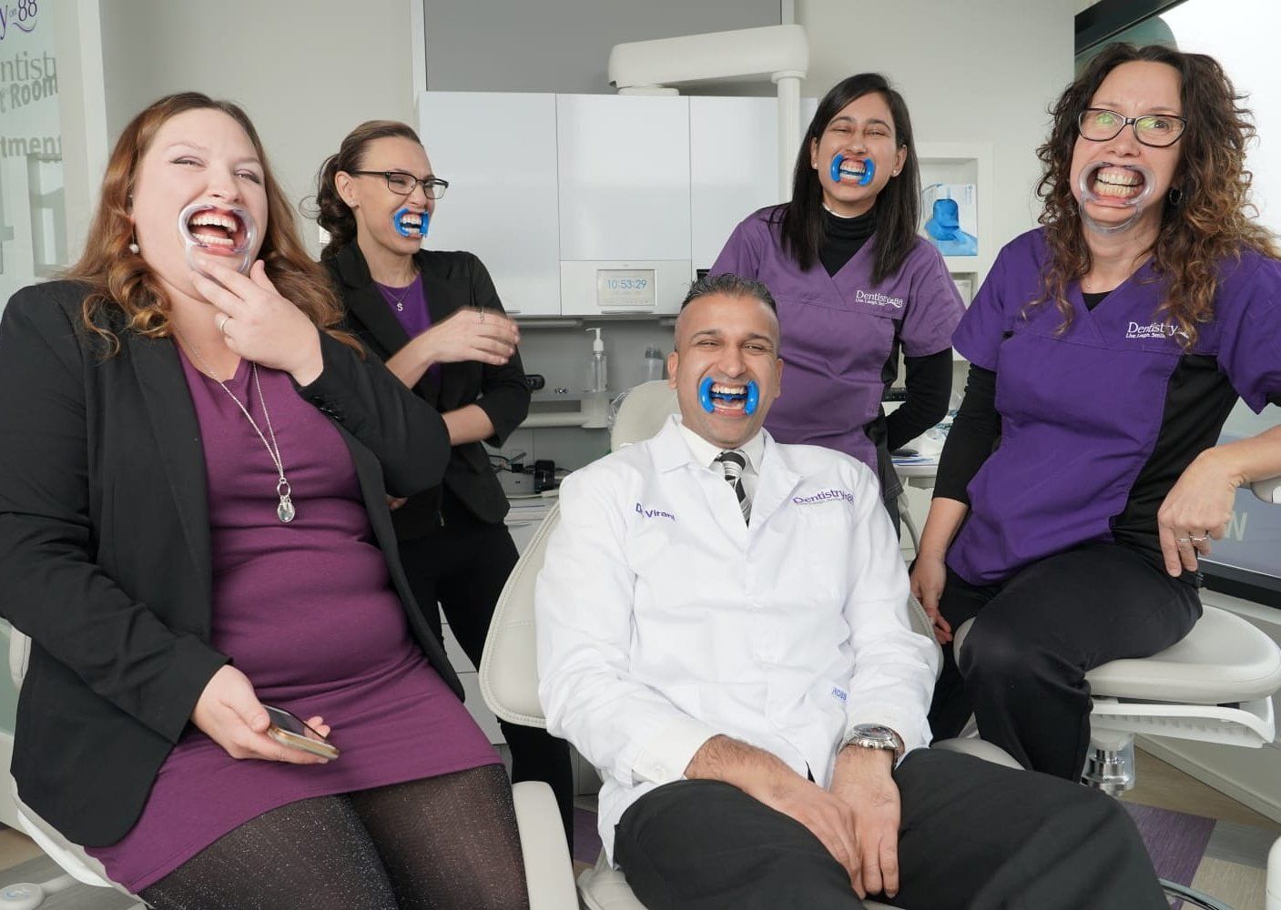 Five smiling dental team members in purple and black uniforms in a bright clinic, one seated in a white coat