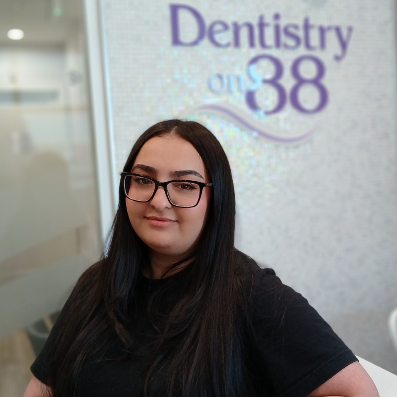 Woman with glasses standing in a dental office, with a “Dentistry on 38” sign behind her.