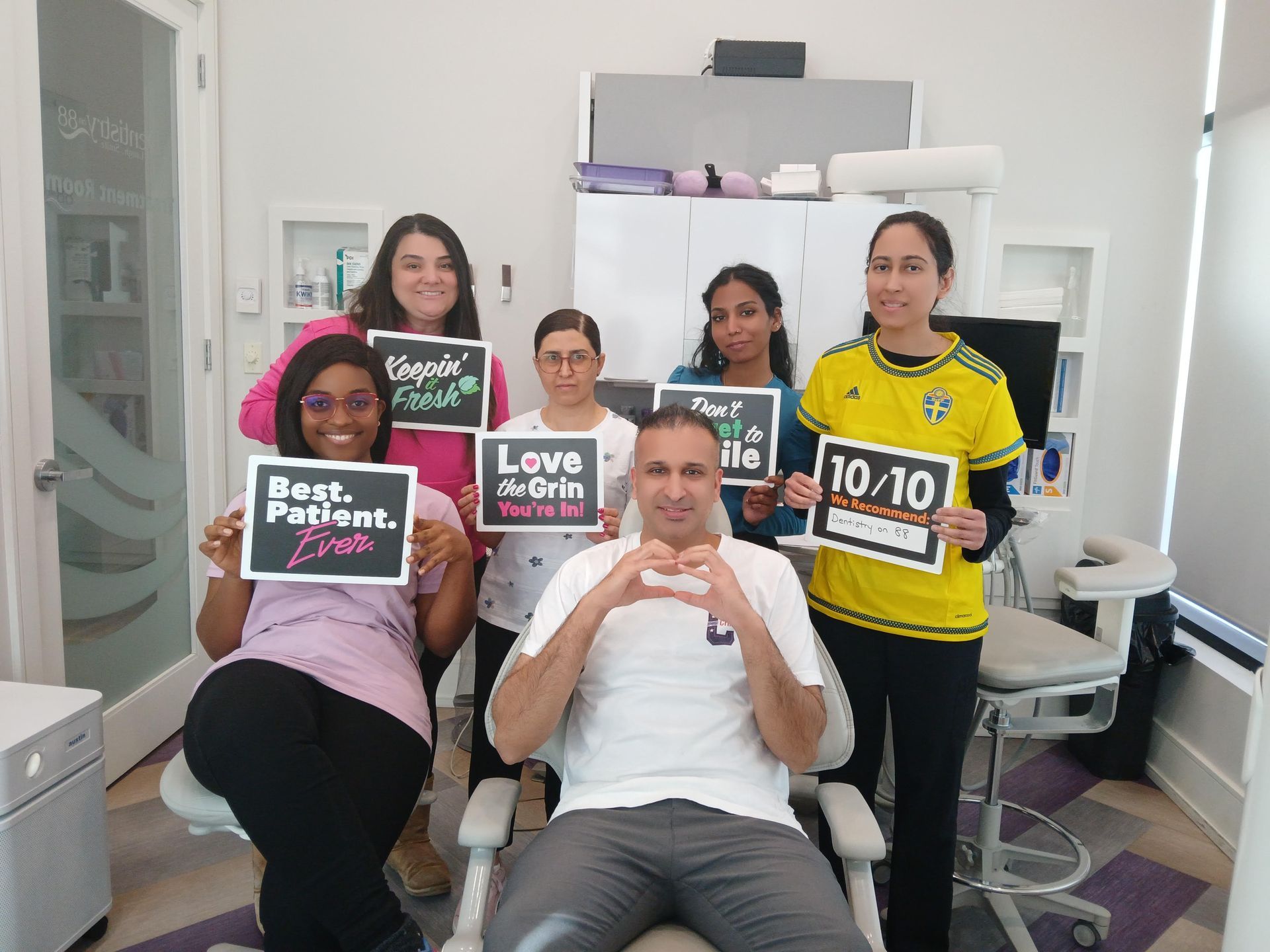 Five people posing in a room, holding race bibs and signs, with one seated on a chair in the center