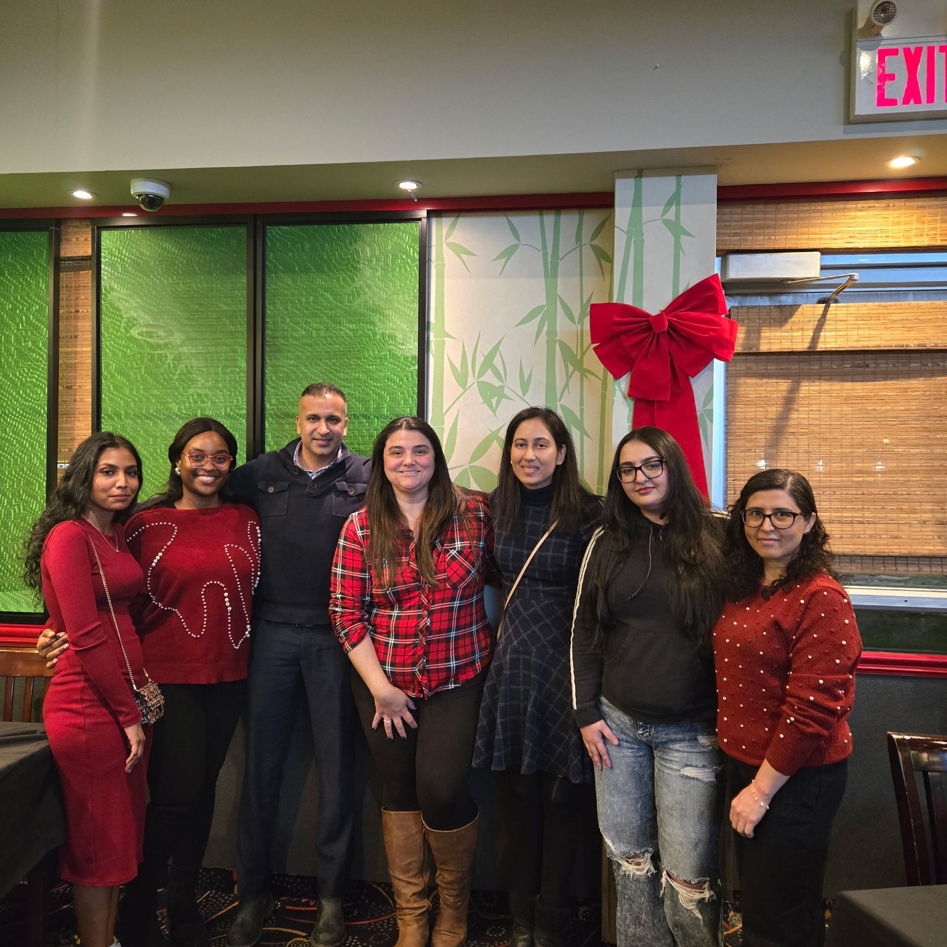 Seven people posing indoors in front of red holiday decorations and a large bow