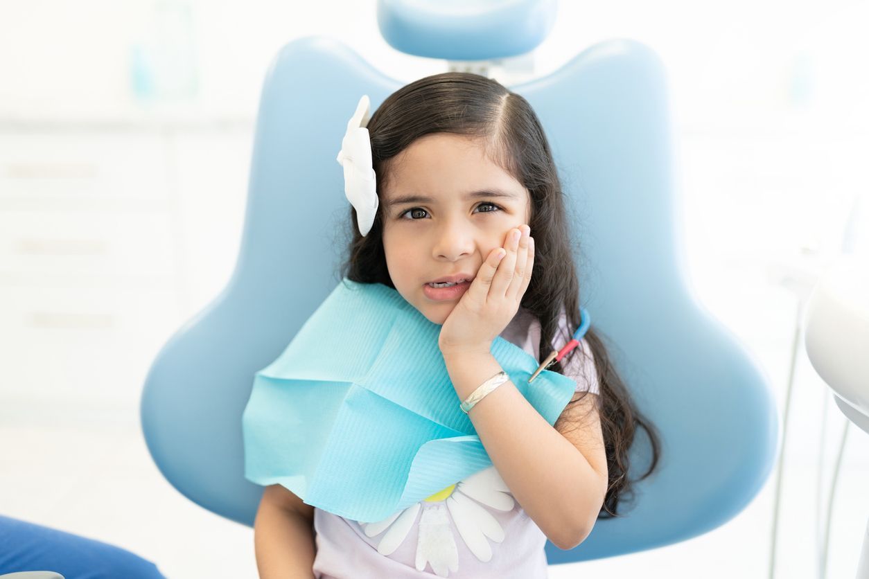 Child sitting in a dental chair, holding a cheek with a blue bib, looking at the camera.