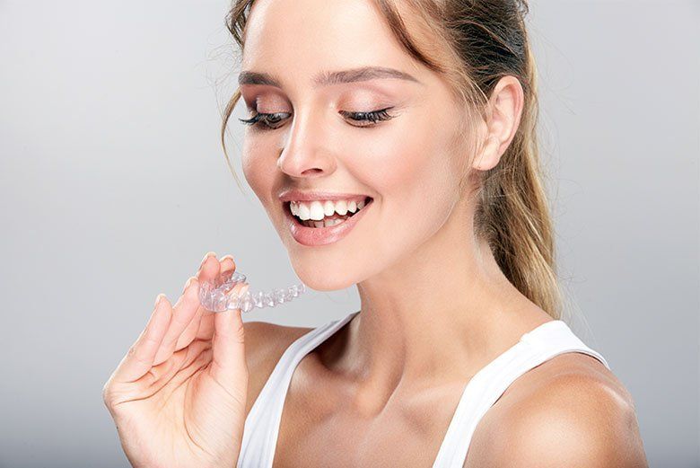 Smiling woman holding a clear dental aligner near her mouth against a light gray background
