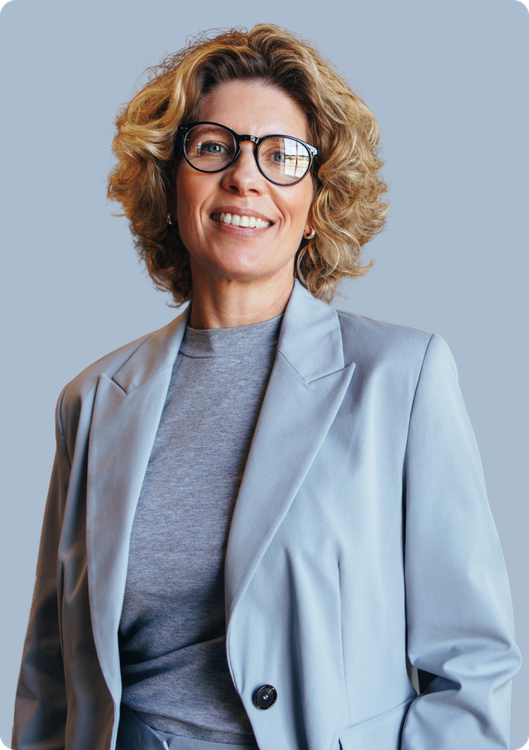 Woman in gray blazer, glasses, and curly hair smiles.