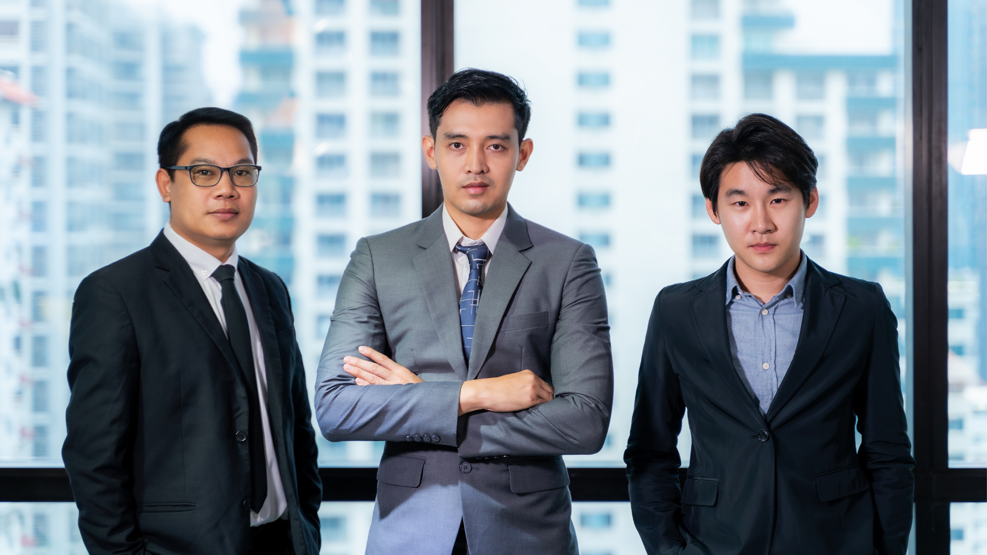 Three business professionals in suits standing in an office with arms crossed.