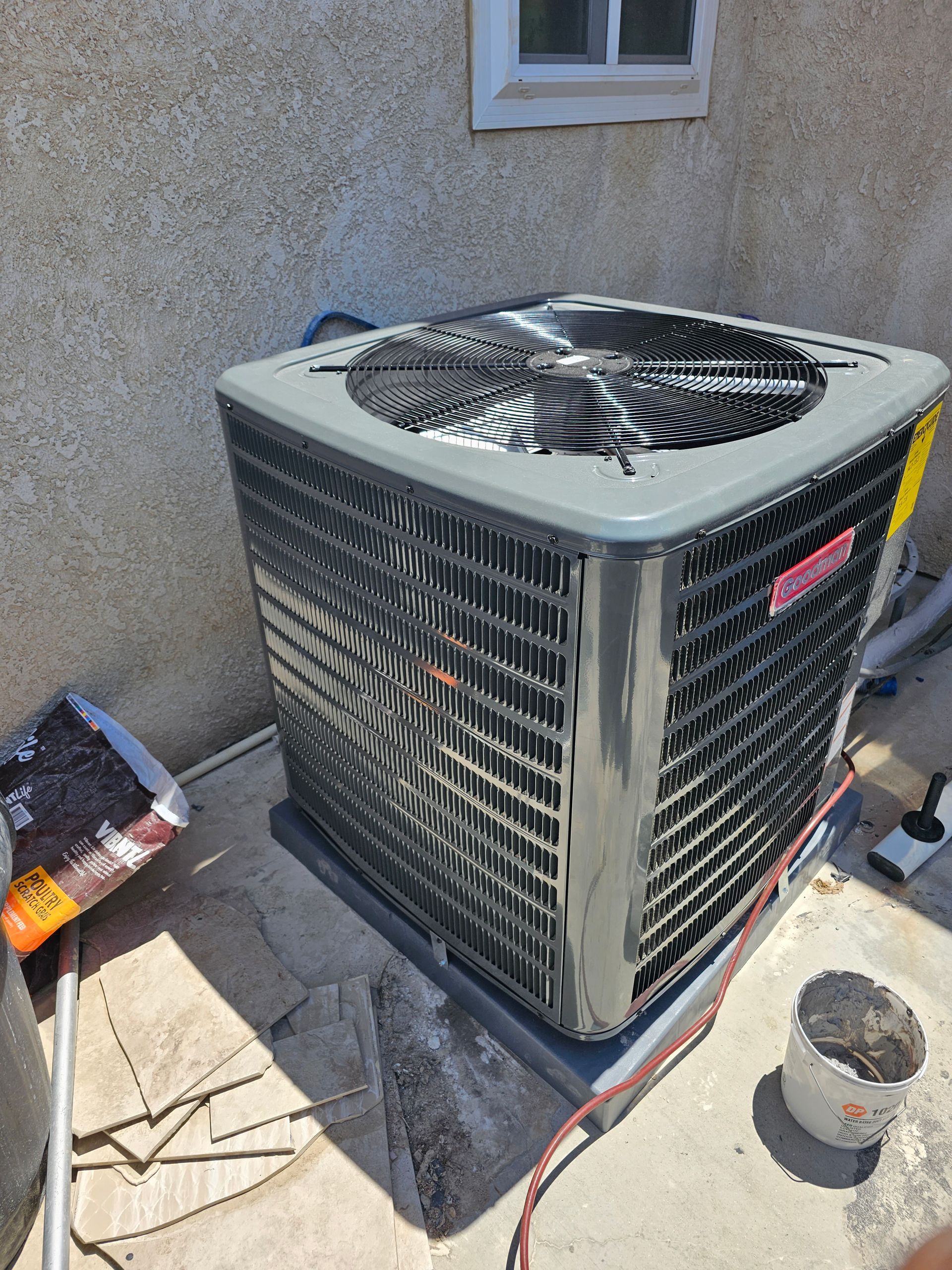 Outdoor HVAC condenser unit beside a stucco wall, with a fan on top and construction debris nearby