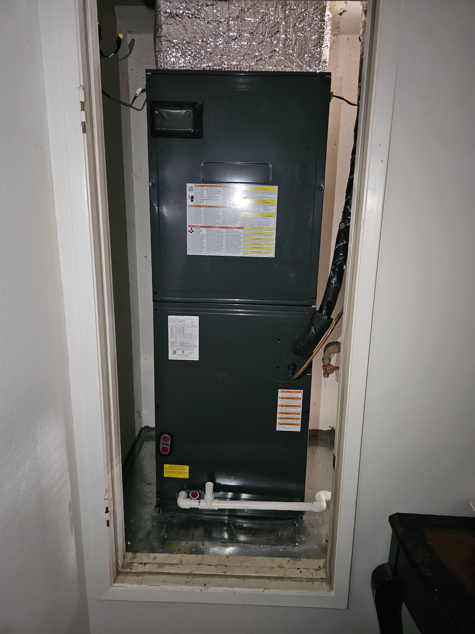 Black HVAC/air handler unit in a narrow closet with exposed ducts and wiring.