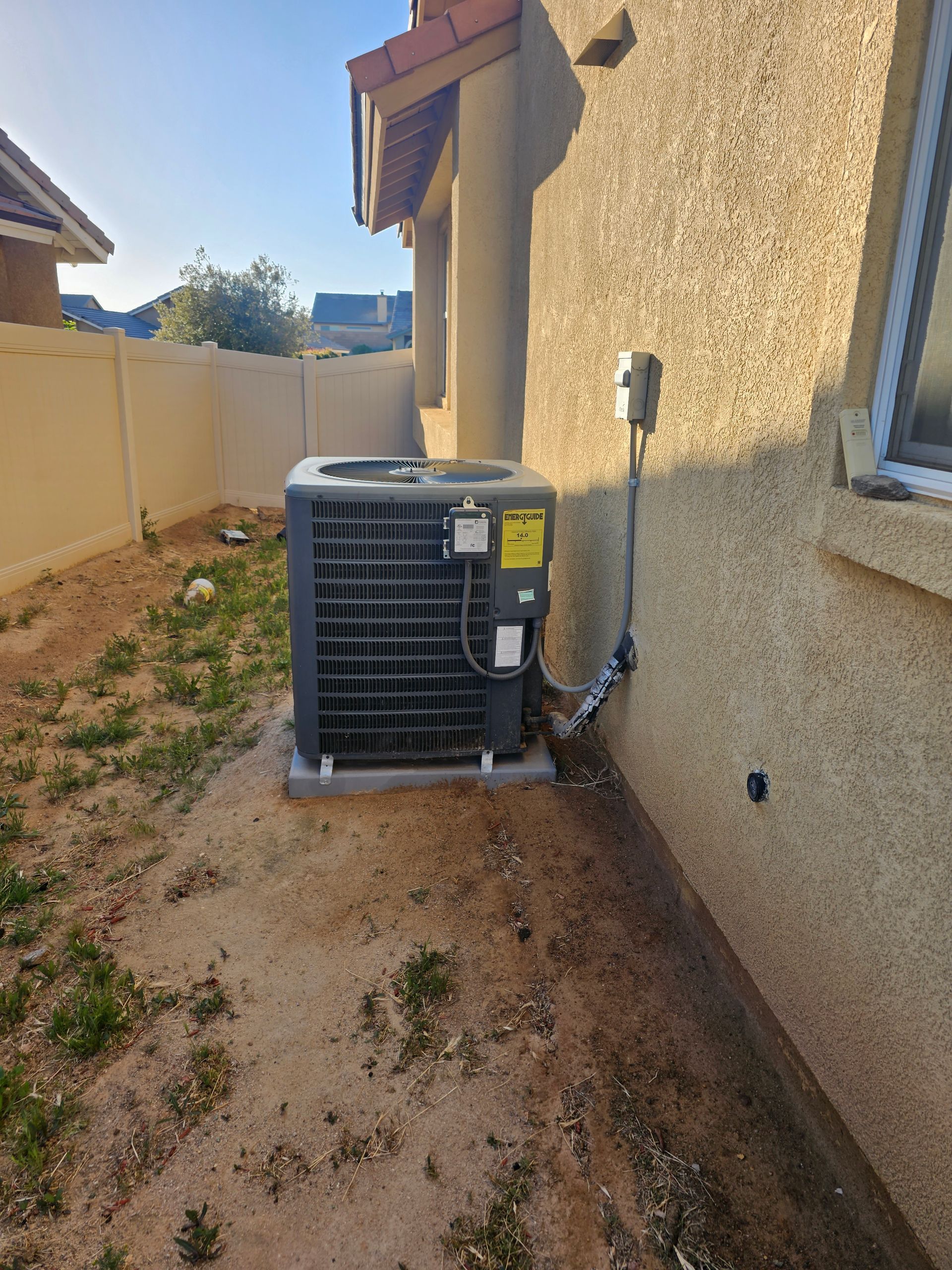 Outdoor air conditioner unit beside a stucco house wall in a narrow dirt side yard