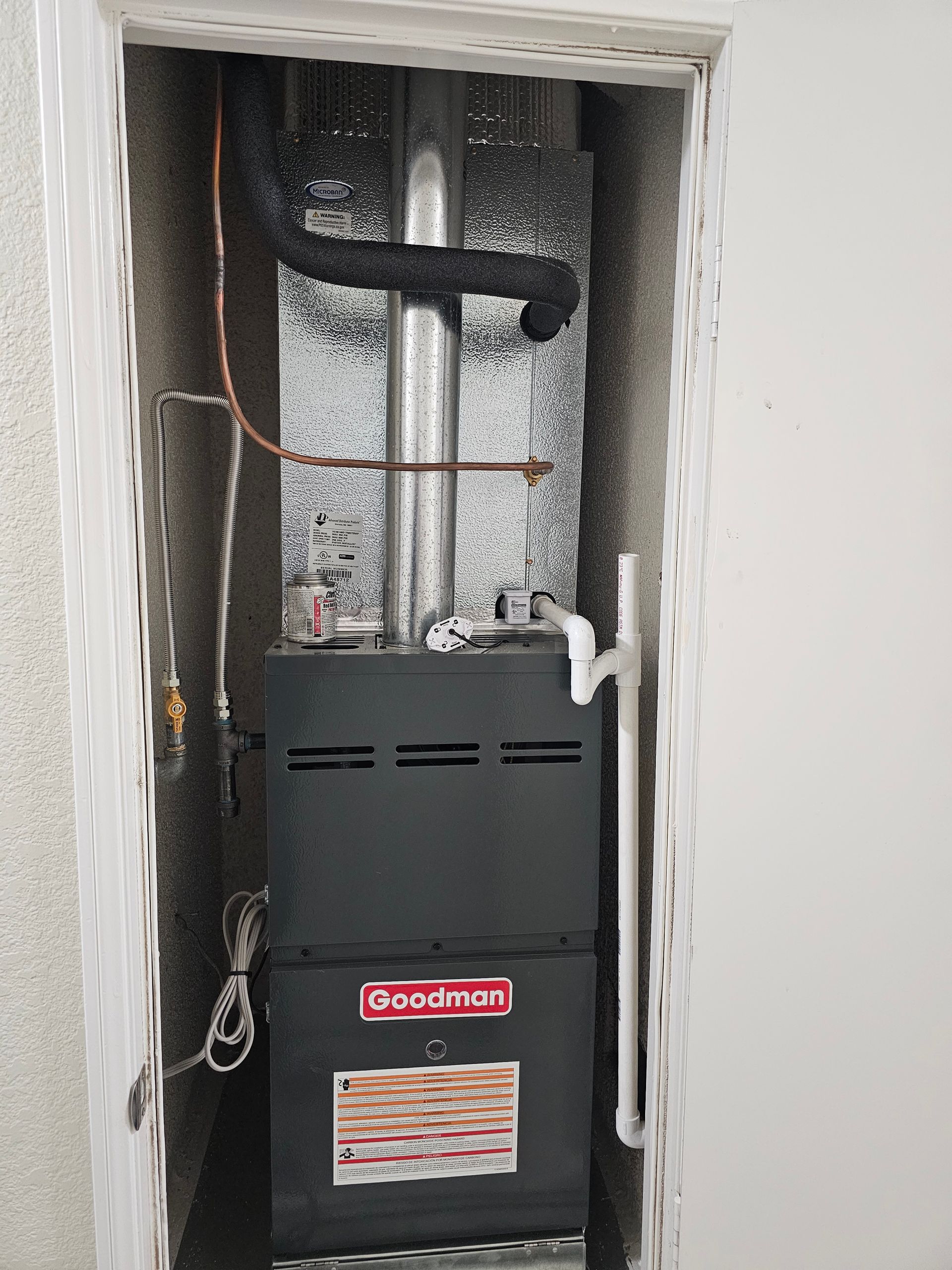 Furnace in a closet with exposed copper pipes and ductwork, against silver insulated backing.