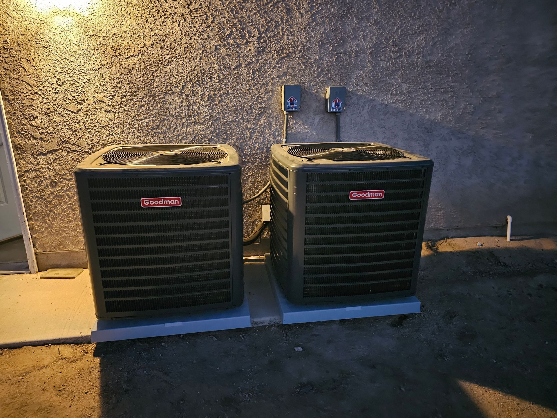 Two outdoor air conditioning units beside a wall at night, lit by a porch light.