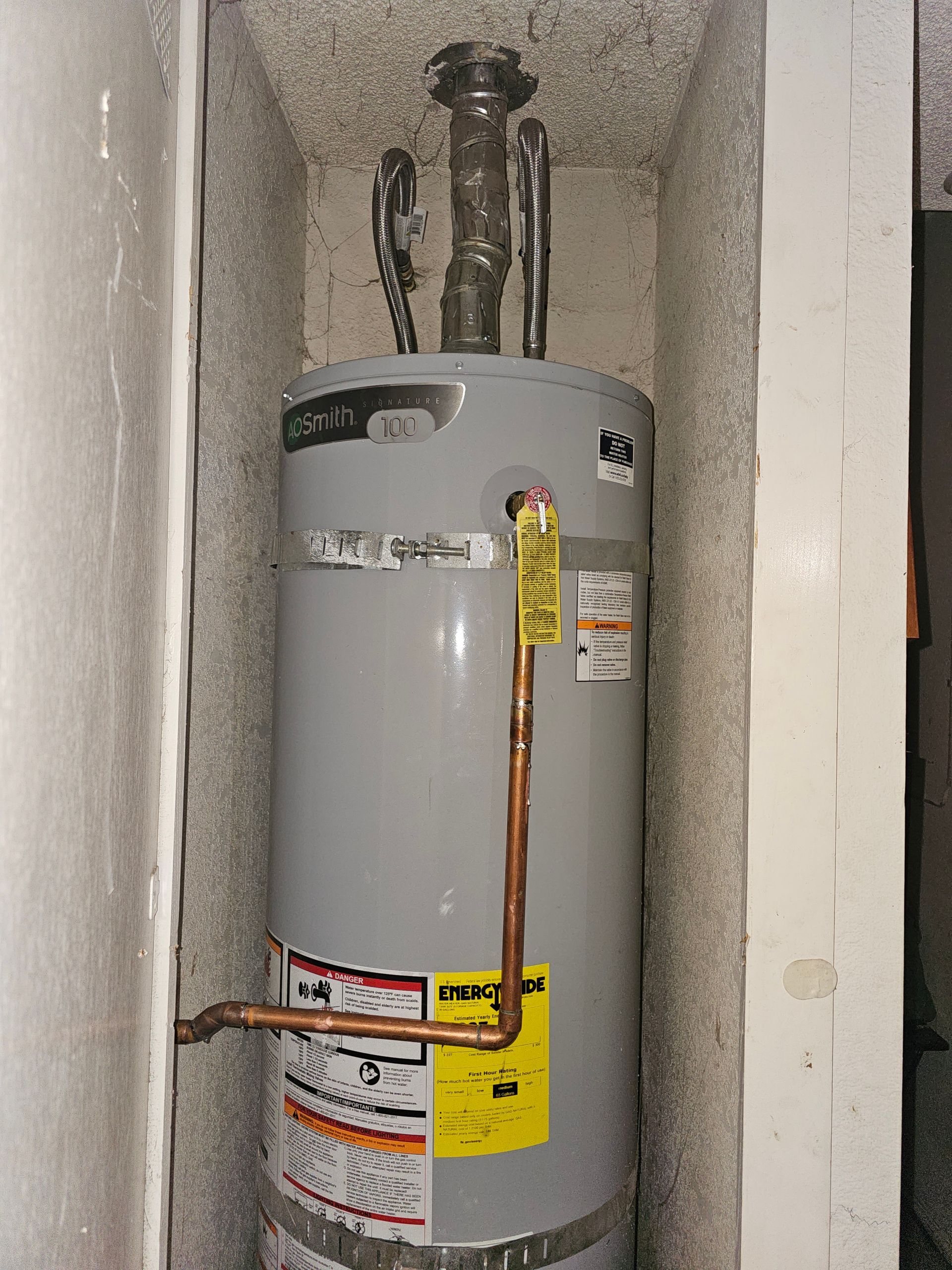 White water heater in a narrow closet with exposed copper piping and yellow safety labels