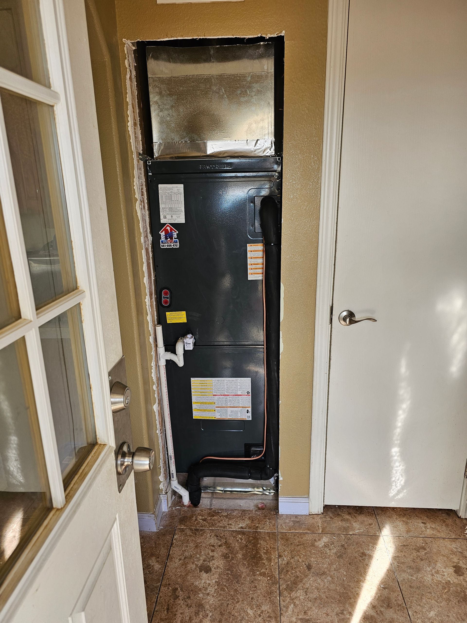 Narrow hallway with a black furnace/utility unit beside a white door and windows, tan carpet, and sunlight on the floor