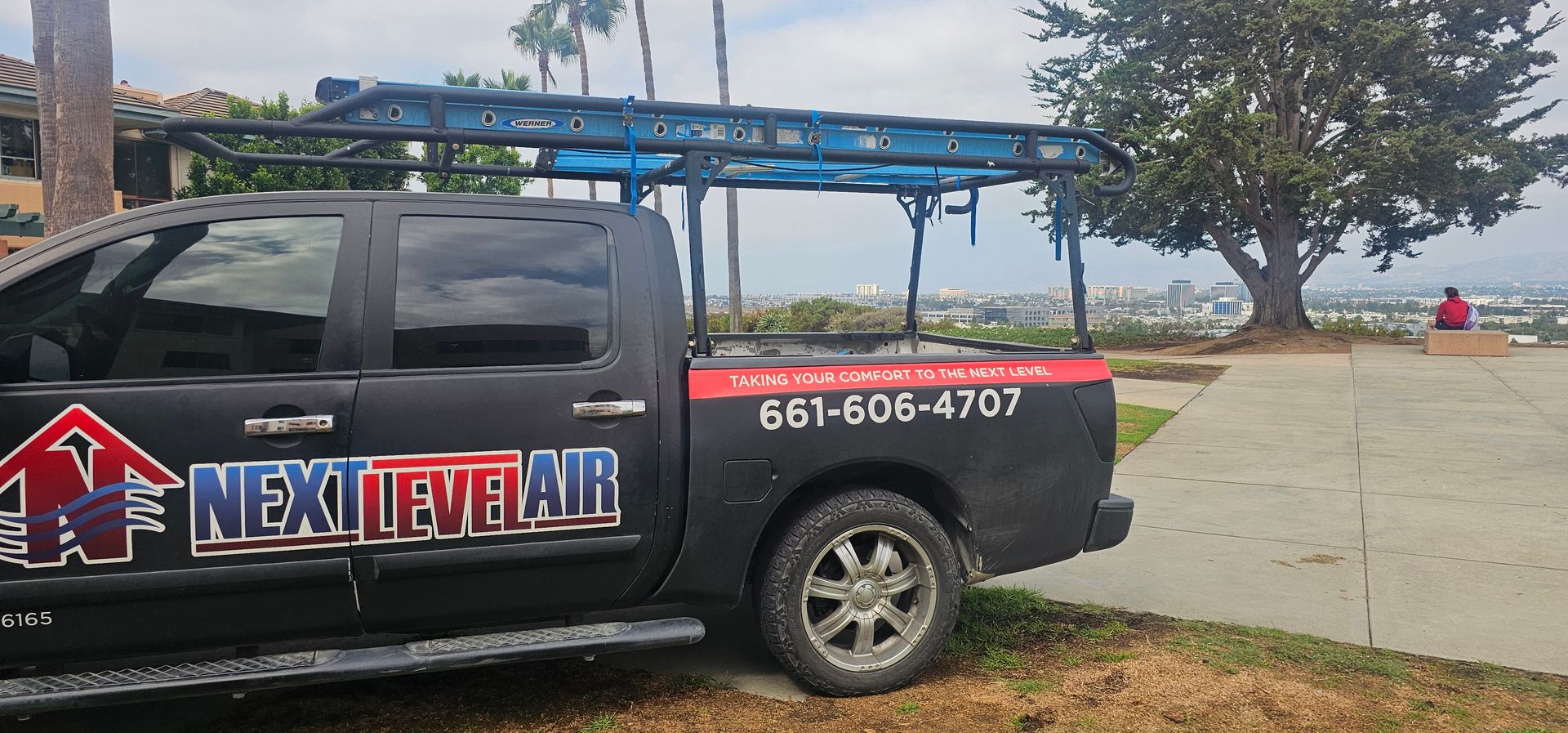 Black pickup with ladder racks and “Next Level Air” logo parked by a waterfront path.