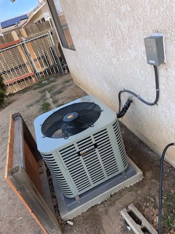 Outdoor air conditioner unit beside a stucco wall with conduit and electrical box, near a fence and wood panel.