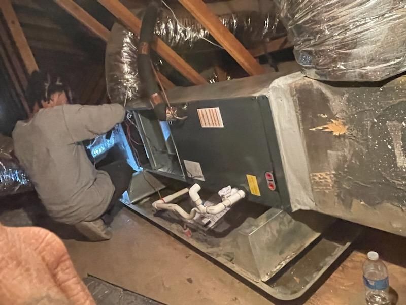 Worker kneeling in a cramped attic beside HVAC ductwork and a furnace unit under exposed rafters.