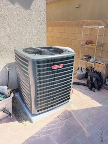 Outdoor HVAC unit beside a tan stucco wall and patio, with a small shelf and equipment nearby
