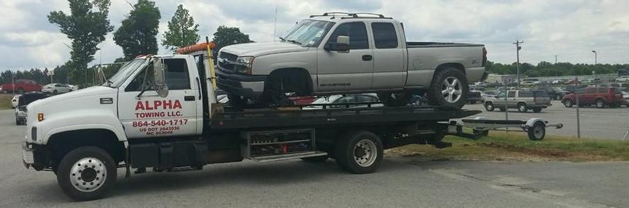 A white Alpha towing truck carrying a silver pickup truck on its flatbed in a parking lot.