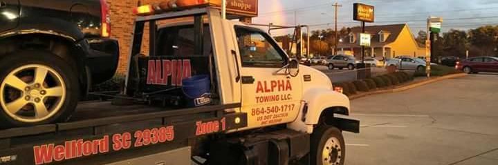 A white tow truck labeled Alpha Towing LLC, parked in a lot in Wellford, SC, carrying a vehicle at sunset.