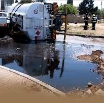A tanker truck is overturned on its side, leaking liquid into a pool on the ground near emergency responders.