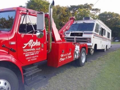 A bright red Alpha Towing truck towing a white recreational vehicle along a roadside on a sunny day.