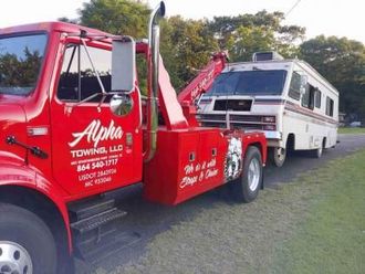 A bright red Alpha Towing truck towing a white recreational vehicle along a roadside on a sunny day.