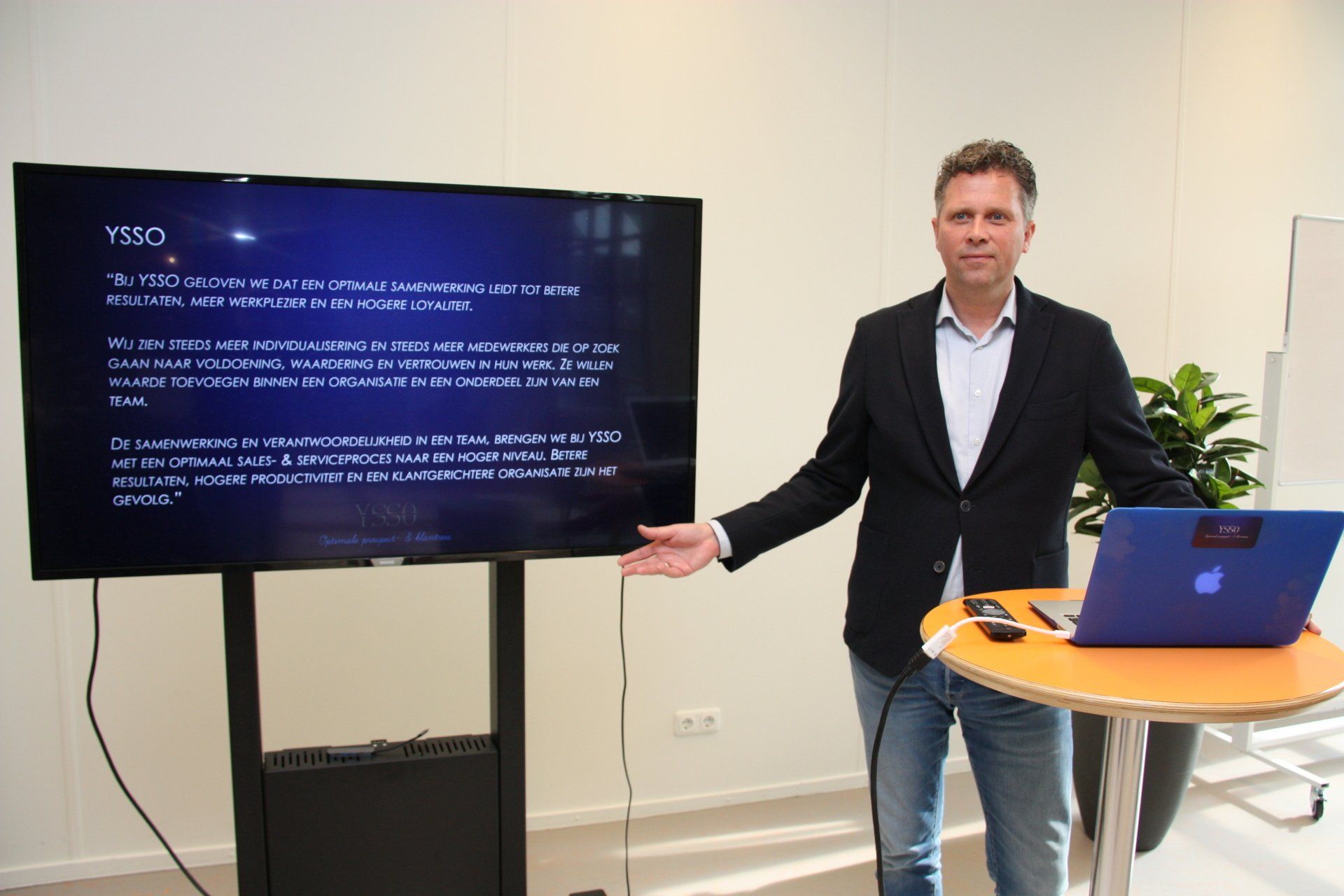 Presenter beside TV screen with slide, standing at a round table with laptop in a bright room