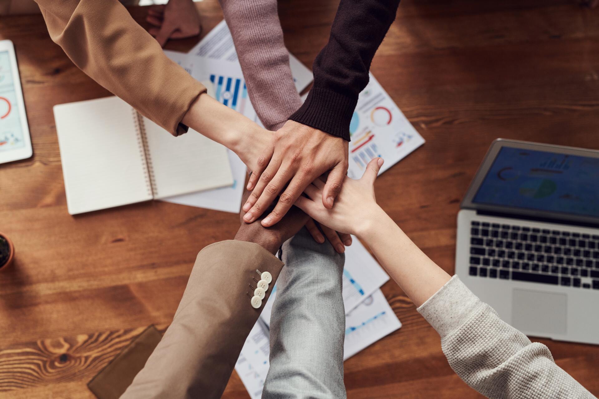 Hands stacked together over a wooden desk with notebooks and a laptop, symbolizing teamwork