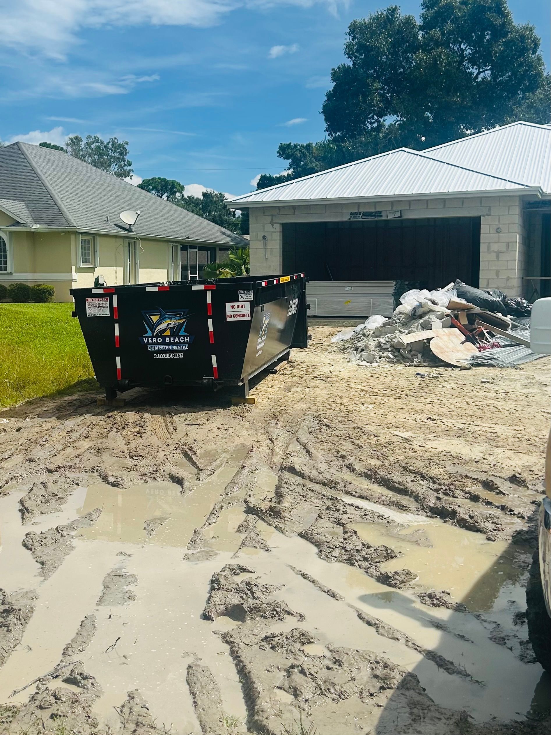 A black construction dumpster sits in a muddy, water-filled yard next to a home with a pile of debris near the garage.