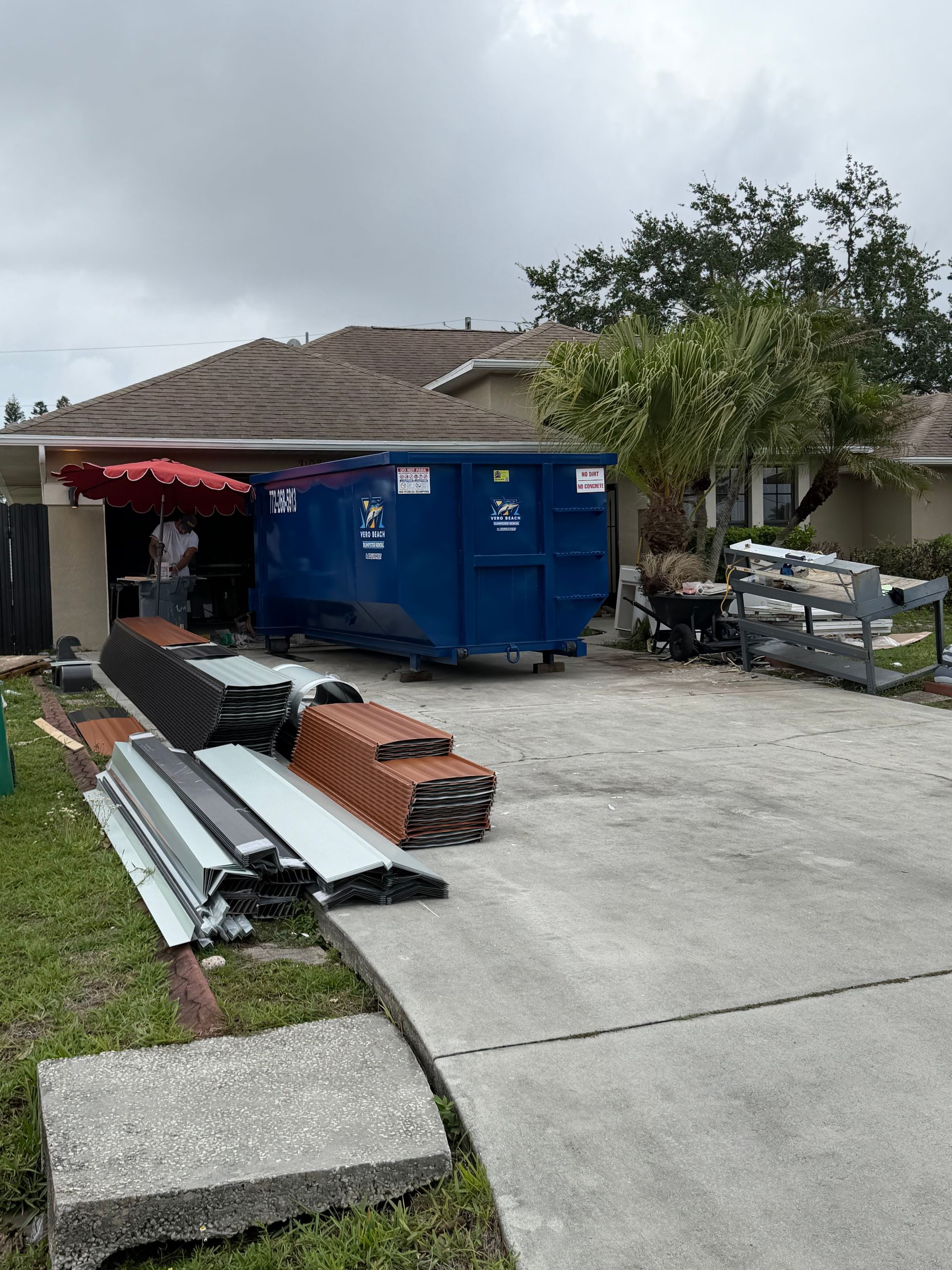 A beige house with a driveway featuring a large black dumpster and stacks of construction materials under a cloudy sky.